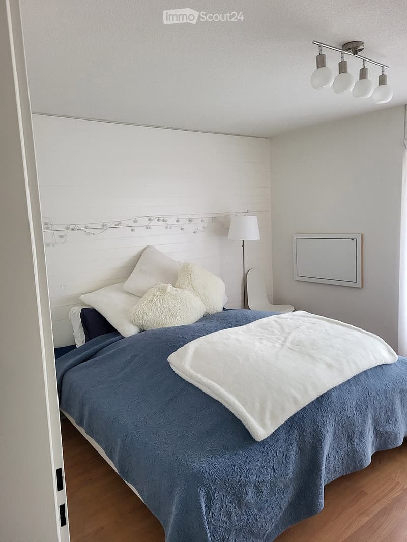 A neatly made bed with blue bedding and white pillows, a white fur throw, a bedside lamp, and a mirror on the wall.