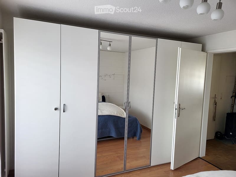 Modern bedroom with large white wardrobe and mirrored doors, reflecting a neatly made bed with blue bedding and wooden flooring.