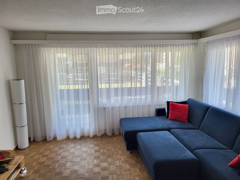 A modern living room with a blue sectional sofa, red throw pillows, large windows with white curtains, and a wooden floor. A white speaker is positioned near the window.