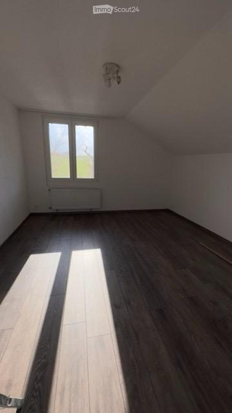 Empty attic room with wooden flooring, white walls, and a single window letting in natural light. The ceiling has a sloped design typical of attic spaces.