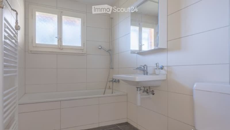 Modern bathroom with white tiled walls and floor, a bathtub, a sink with a mirror, and a toilet.