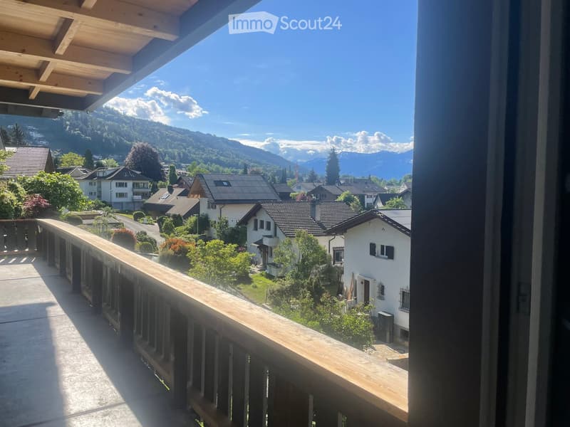 Balcony overlooking a scenic town with houses, greenery, and distant mountains under a clear blue sky.
