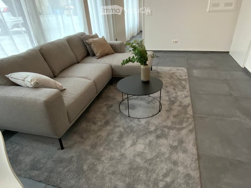 Modern living room with a grey sectional sofa, a black coffee table, a large grey rug, and a potted plant on the coffee table. The room features large windows with sheer curtains and a grey tiled floor.