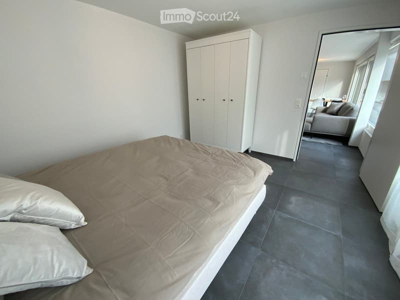 Modern bedroom with a large bed, white bedding, and a white wardrobe. The room has dark tiled flooring and large windows with blinds.
