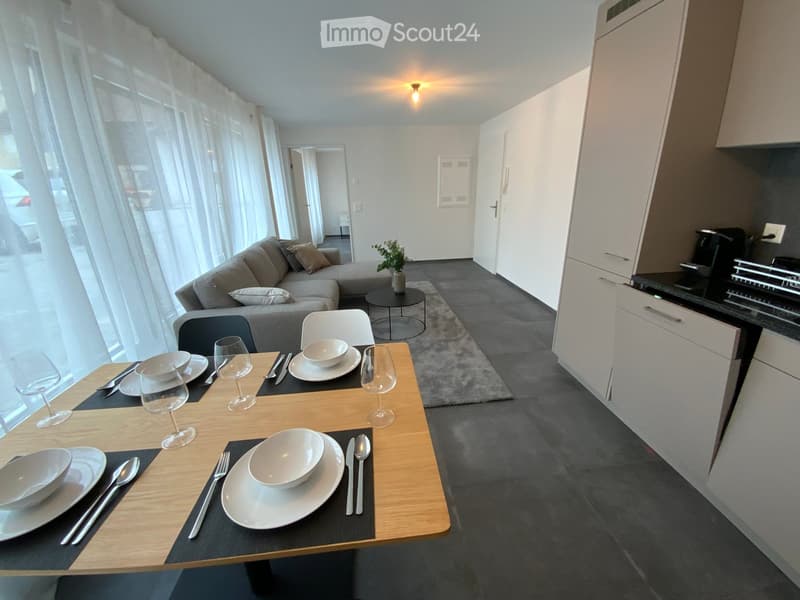 Modern open-plan living and dining area with a grey sofa, dining table set for two, white cabinets, and a small kitchen area.