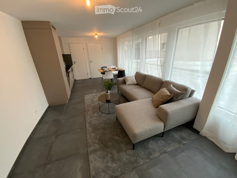 Modern living room with a grey sectional sofa, coffee table, dining area with chairs, and large windows with sheer curtains.