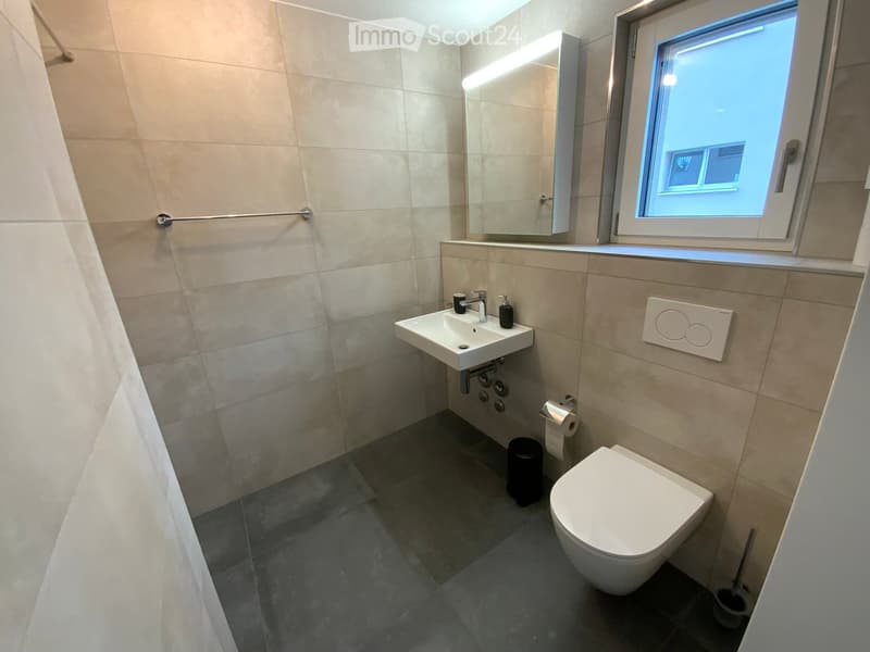 Modern bathroom with a toilet, sink, mirror, and window. Features beige tiles, a dark grey floor, and a sleek design.
