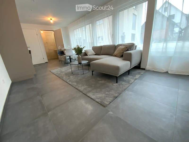 Modern living room with a grey sectional sofa, ottoman, coffee table, and a small dining area with chairs and a table.