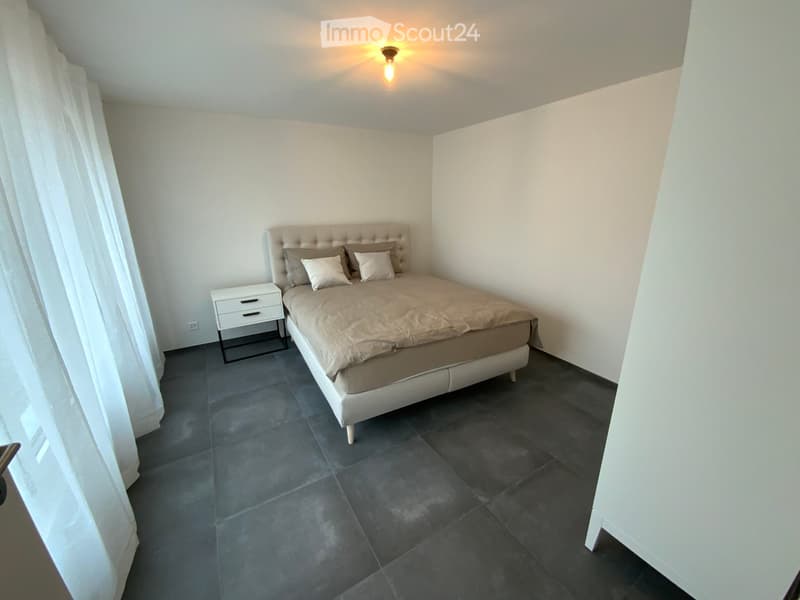 Modern bedroom with a large bed, nightstand, and pillows, featuring dark tiled flooring and white walls.