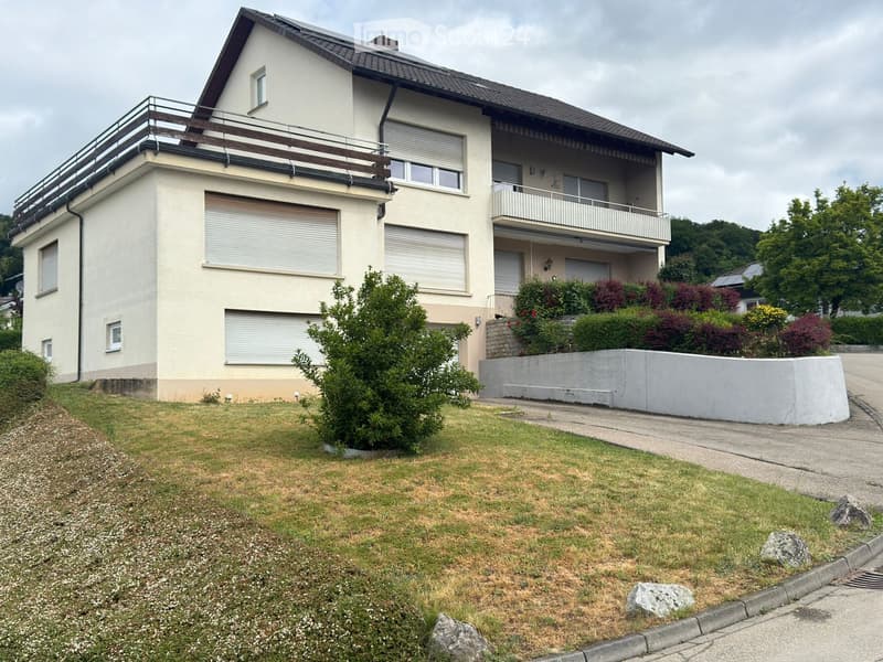 Two-story house with beige exterior, multiple windows, balconies, and surrounding greenery.