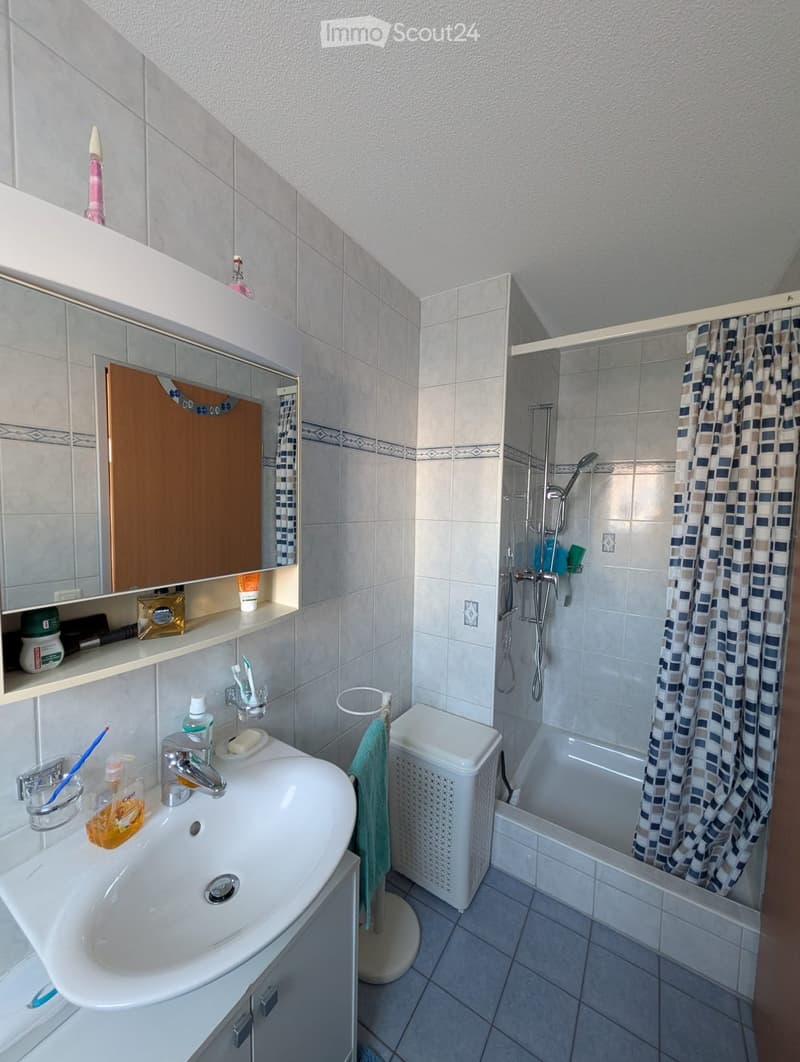 white bathroom with tiled walls and floor, vanity with sink, mirror, and a shelf, shower with showerhead and shower curtain, white bathtub, towel rack with a towel, white trash bin.