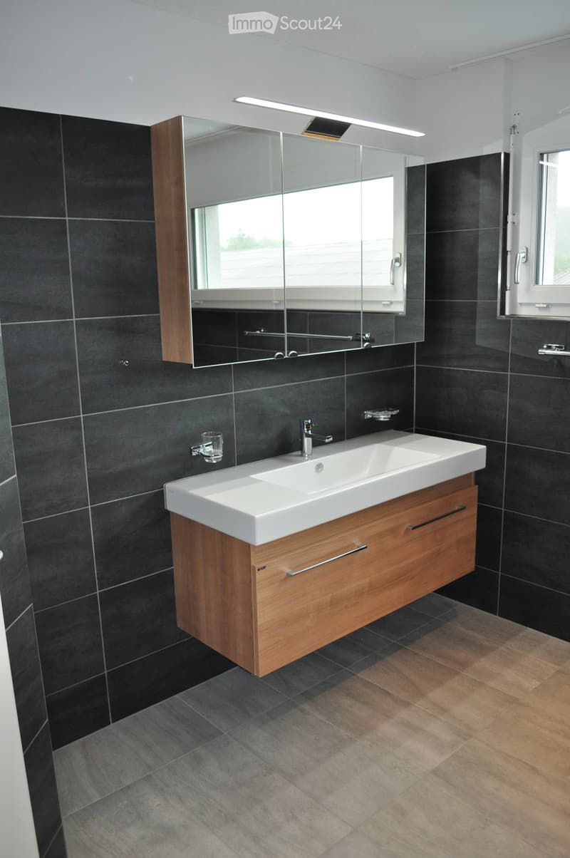 Modern bathroom with dark tiled walls, a white sink with wooden cabinet, a large mirror, and a window providing natural light.