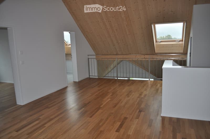 An empty attic space with wooden flooring, white walls, and a metal railing leading to a staircase. The ceiling is slanted with wooden beams and a skylight window allowing natural light.