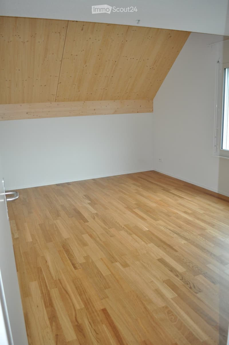 Empty room with light wooden flooring and white walls, featuring a slanted ceiling with wooden paneling.