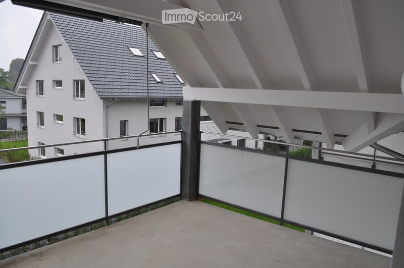 Modern balcony with white walls, metal railing, and a view of a contemporary house with a grey tiled roof and multiple windows.