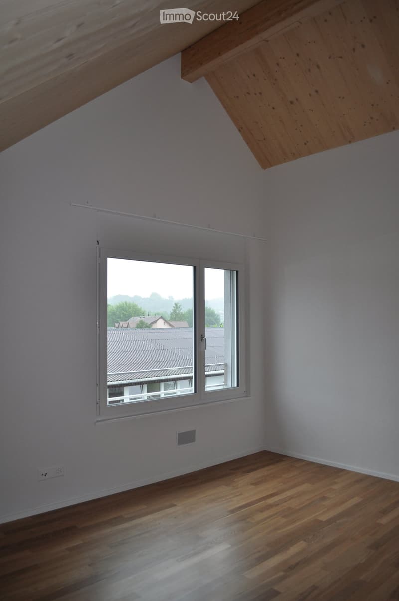 Empty room with wooden flooring, white walls, and a large window offering a view of neighboring rooftops and trees.
