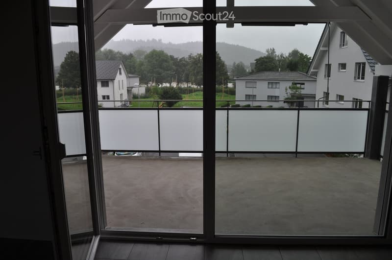 Balcony with glass railing, concrete floor, overlooking a residential area with houses and greenery.