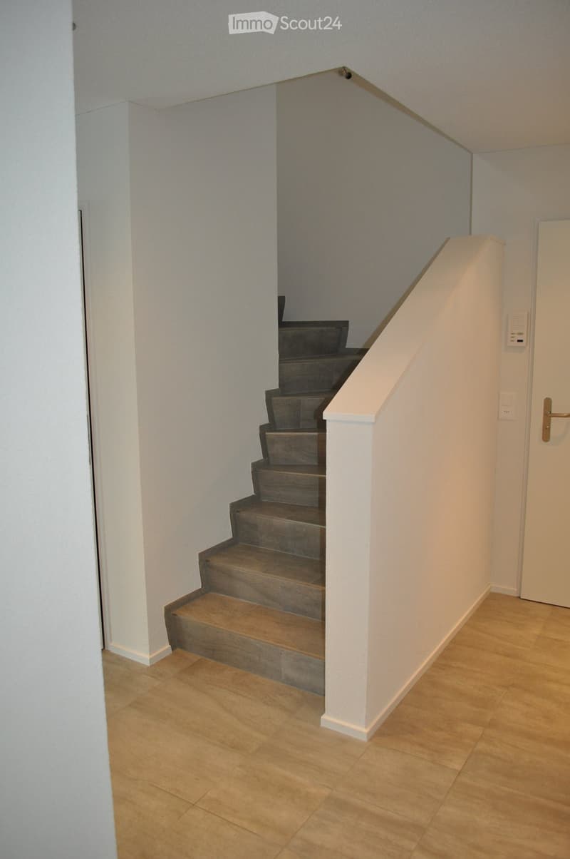 A modern hallway with wooden flooring, a staircase with gray steps, white walls, and a white door on the right side.