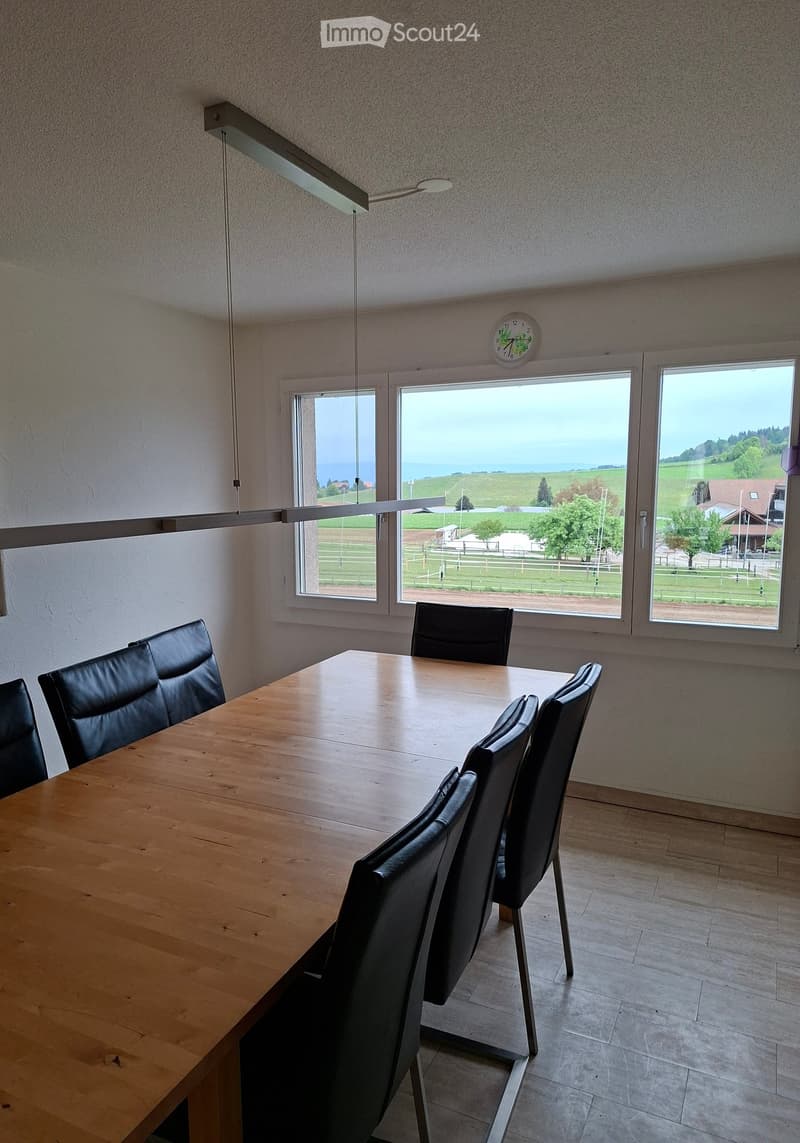Bright room with large windows overlooking a scenic landscape, featuring a wooden table and black leather chairs.
