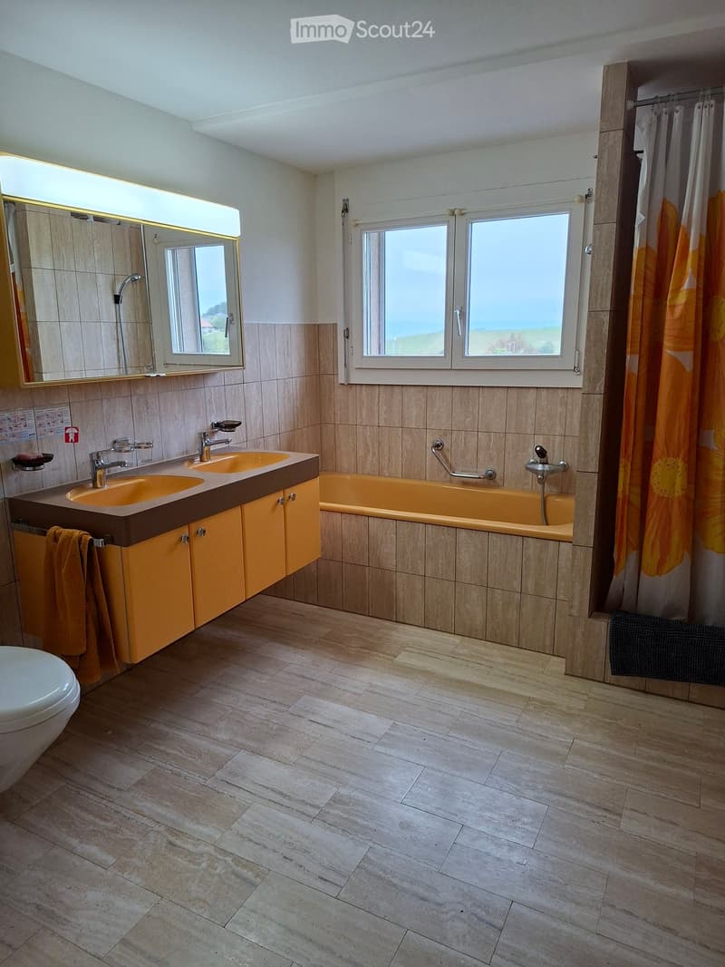 Modern bathroom with double sink, bathtub, large mirror, and natural light from a window. Features light wood flooring and beige tiles.