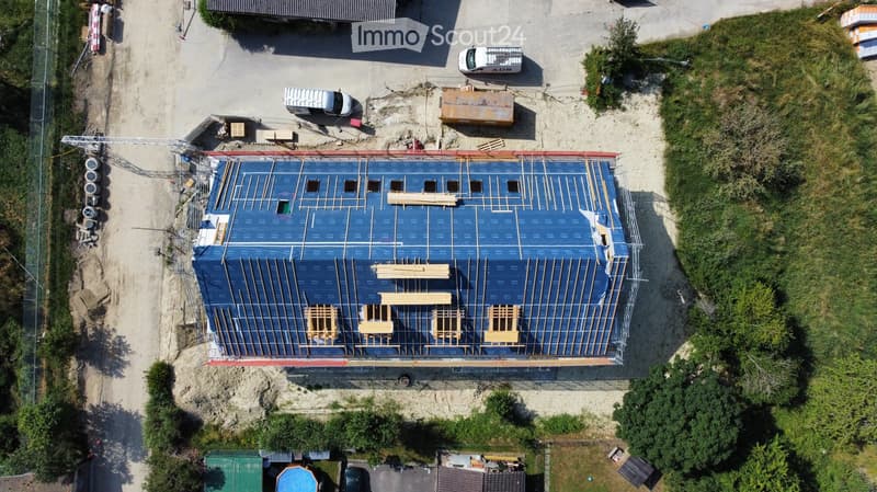 Aerial view of a building under construction with a blue tarp-covered roof and exposed wooden beams. Surrounding area includes construction materials, vehicles, and greenery.