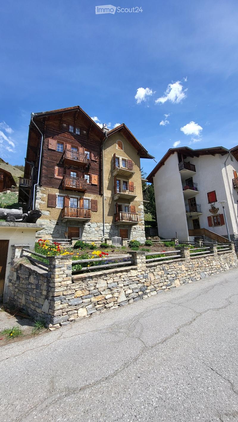 A multi-story residential building with wooden balconies and stone facades, surrounded by a stone wall and colorful flowers.
