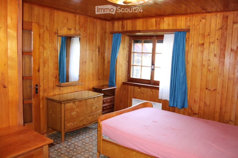 Cozy wooden-paneled bedroom with a single bed, dresser, mirror, and nightstand. Features wooden walls, a window with blue curtains, and a patterned tile floor.