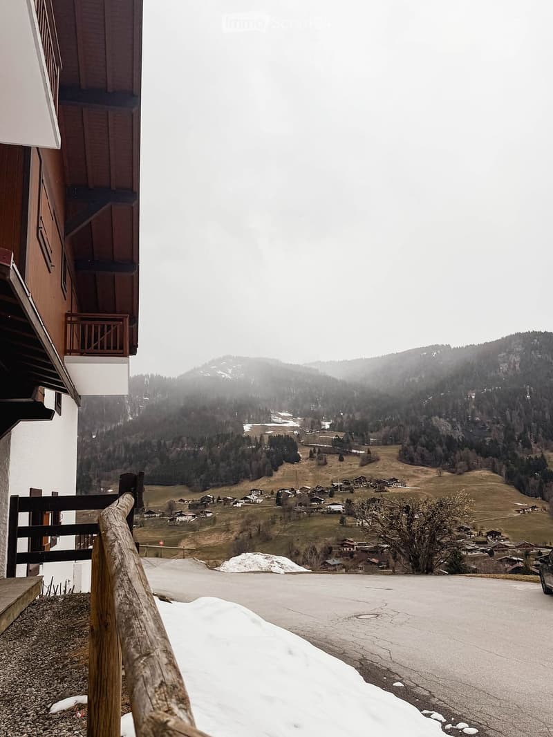 Balcony overlooking a snowy village and forested hills, with wooden railing and a view of the road below.