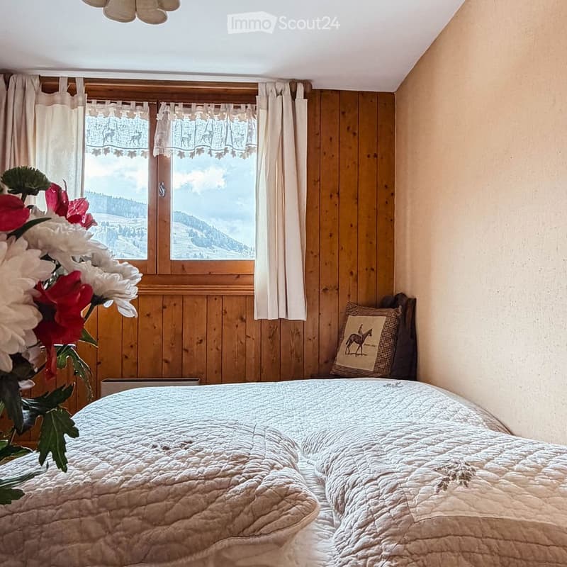 Cozy bedroom with wooden walls, large window offering a scenic mountain view, a neatly made bed with a quilt, and a decorative pillow on a chair.