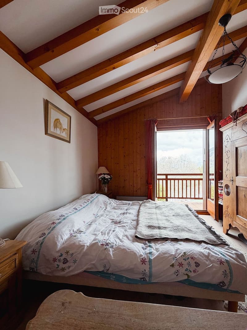 Cozy attic bedroom with wooden beams, a large bed, nightstand, lamp, picture frame, and a balcony overlooking a scenic view.