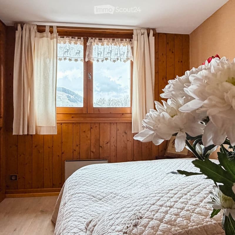 Cozy bedroom with wooden walls, large window with curtains, and a neatly made bed with a white quilt. A vase of white flowers is placed on a surface near the window.