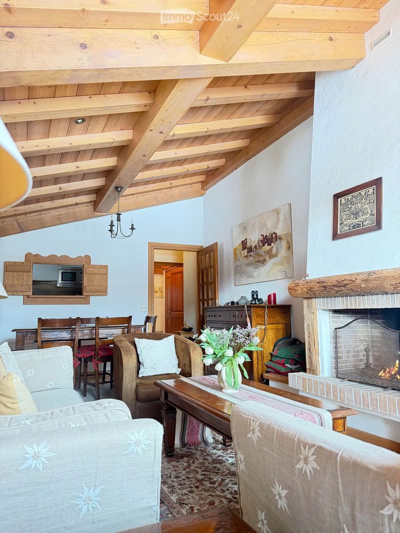 Cozy living room with exposed wooden beams, a fireplace, a sofa, coffee table, dining area, and artwork on the walls.
