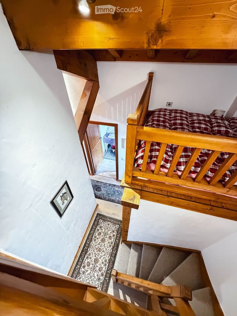 Wooden staircase with a wooden railing and a red and white checkered blanket on the landing. The walls are white, and there are patterned rugs on the stairs. A framed picture is on the wall, and a doorway leads to another room.