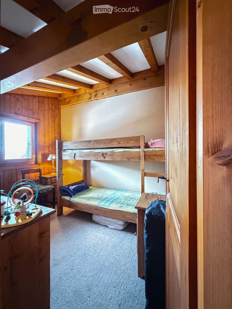 Cozy attic bedroom with wooden bunk bed, nightstand, chair, and small table lamp. The room features exposed wooden beams and a window letting in natural light.