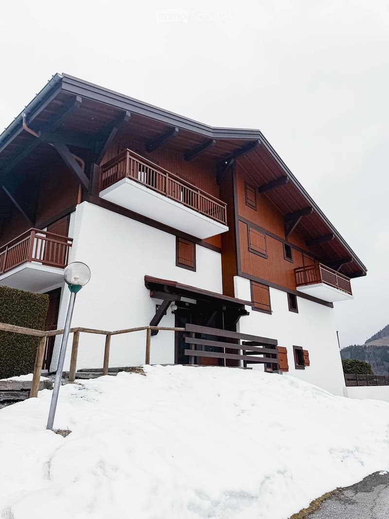 A two-story wooden house with white walls, multiple balconies, and wooden shutters, surrounded by snow.