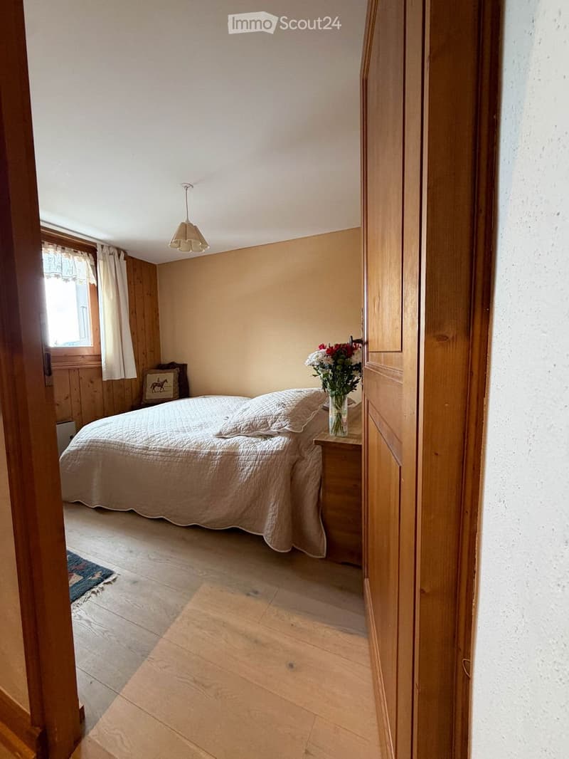 Cozy bedroom with wooden walls, a bed with a light-colored duvet, a nightstand with a vase of flowers, a picture frame on the wall, and a small rug on the floor.