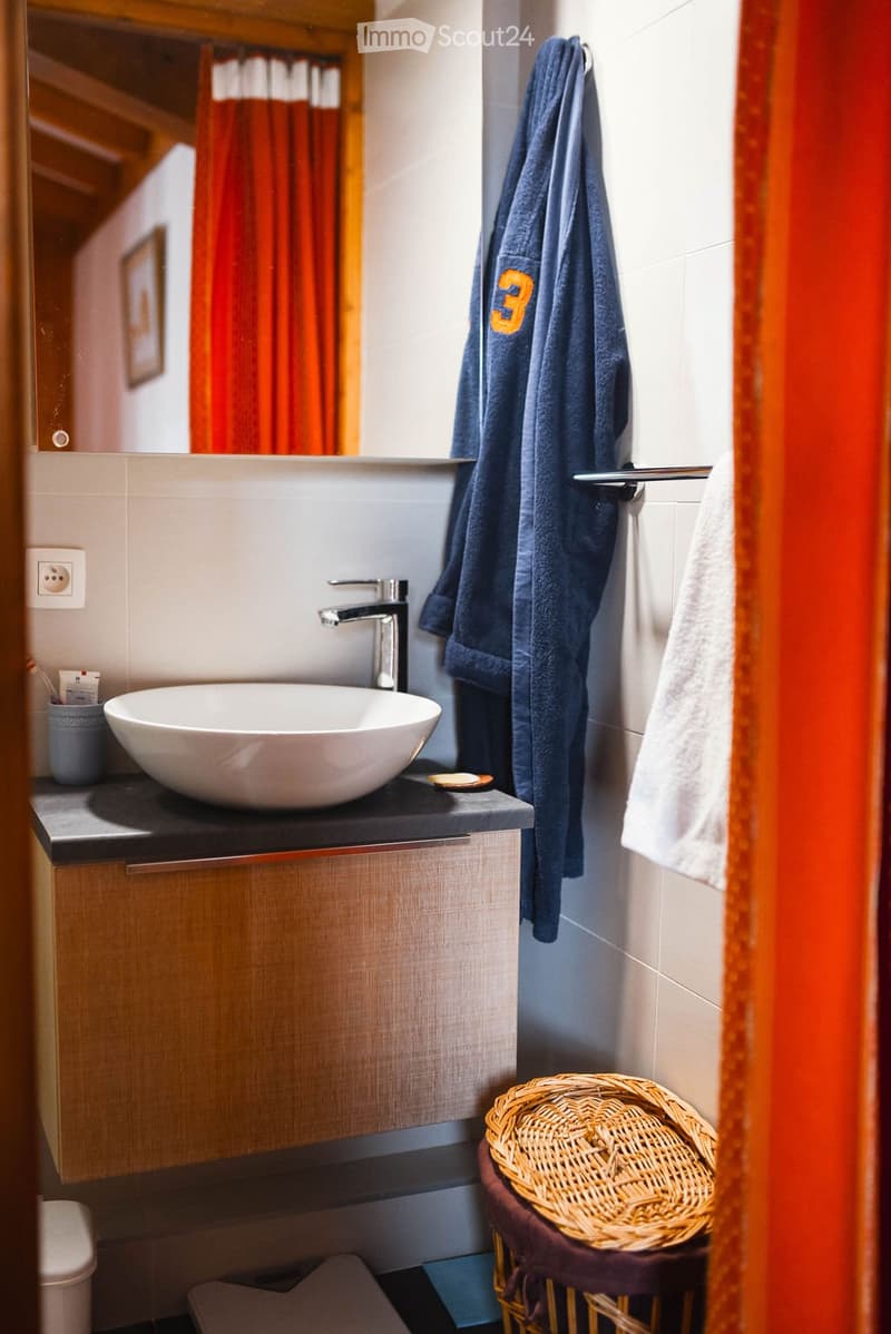 Modern bathroom with a white sink, sleek faucet, wooden cabinet, and a wicker basket on a small table. A blue towel with the number "3" is hanging on the wall.