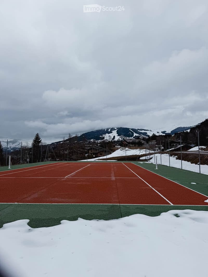 An outdoor tennis court with red and green surfaces, surrounded by a fence and snow-covered ground, with mountains and houses in the background.