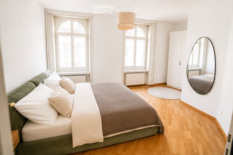 Bright bedroom with a large bed, green sofa, white pillows, a round mirror, a wardrobe, and a pendant light hanging from the ceiling.