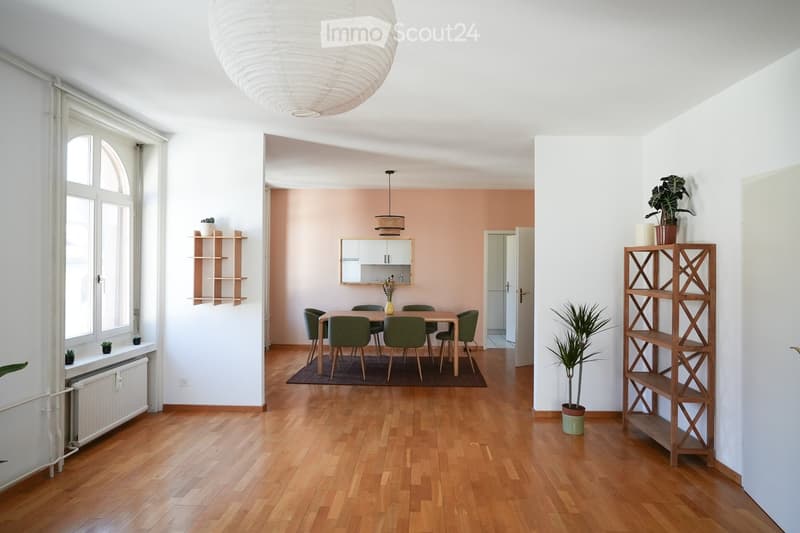 Bright, open-plan living and dining area with wooden flooring, large windows, a round pendant light, a dining table with six chairs, shelves with plants, and a small kitchen area with cabinets and a mirror.