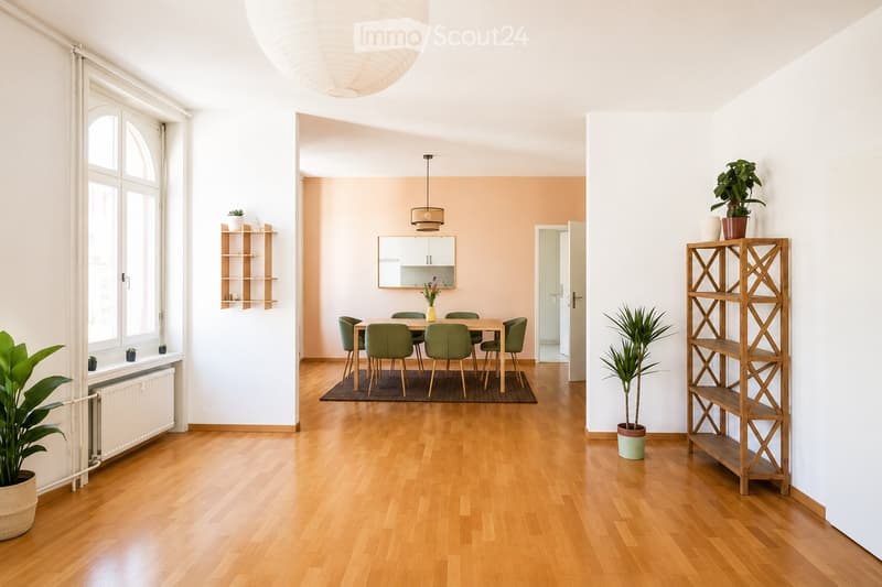 Bright, open-plan living and dining area with wooden flooring, large windows, and modern furniture including a dining table, chairs, shelves, and potted plants.