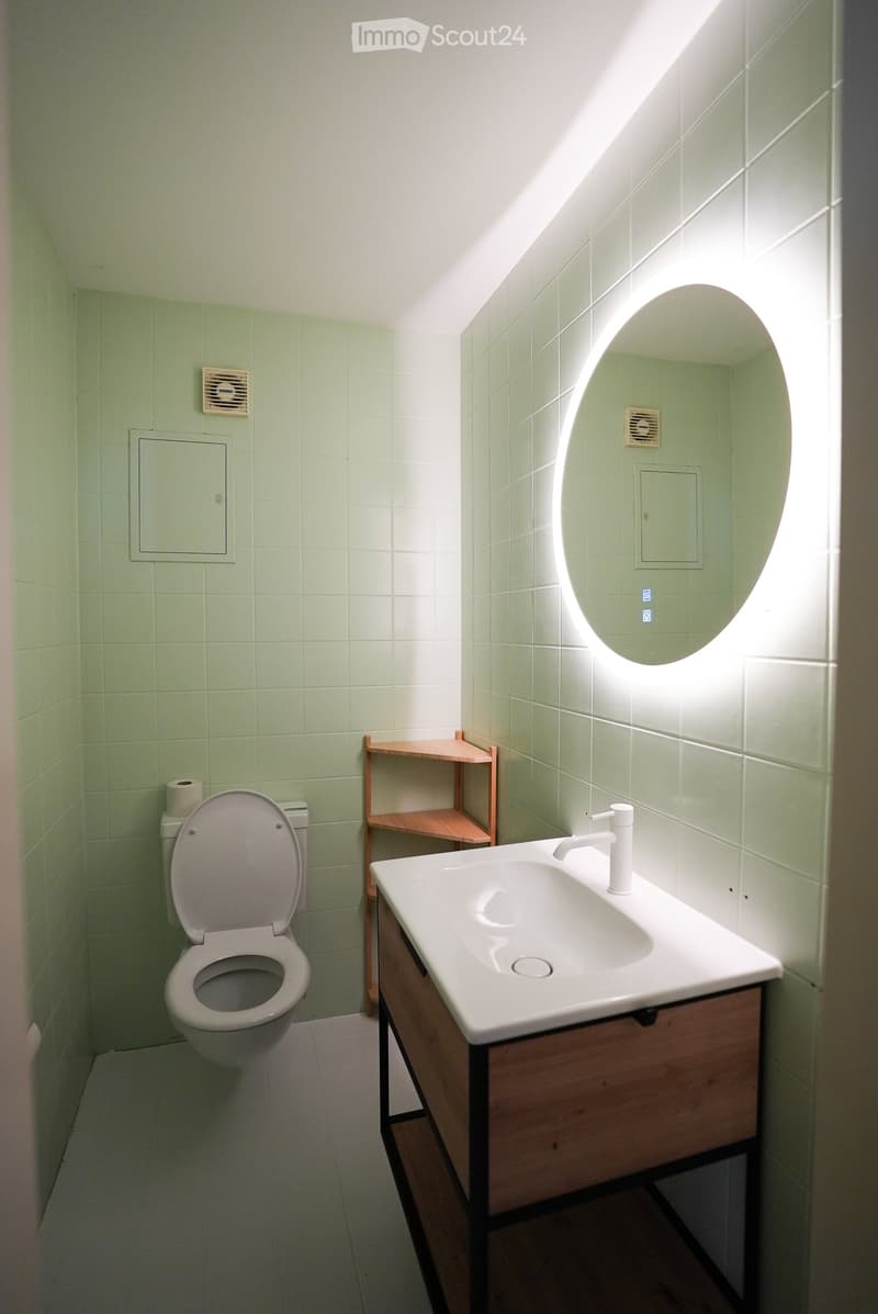 Modern bathroom with light green tiled walls, white sink with wooden cabinet, round illuminated mirror, toilet, and small wooden shelf.