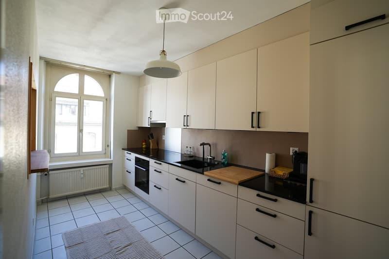 Modern kitchen with white cabinets, black countertops, and a large window. Features include a sink, oven, coffee maker, and various small appliances.