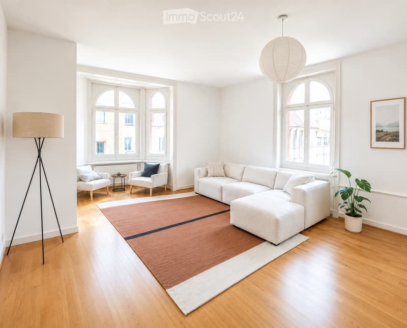 Bright living room with large arched windows, white walls, light wood flooring, a sectional sofa with an ottoman, two armchairs, a small side table, a floor lamp, a potted plant, and a picture frame on the wall.