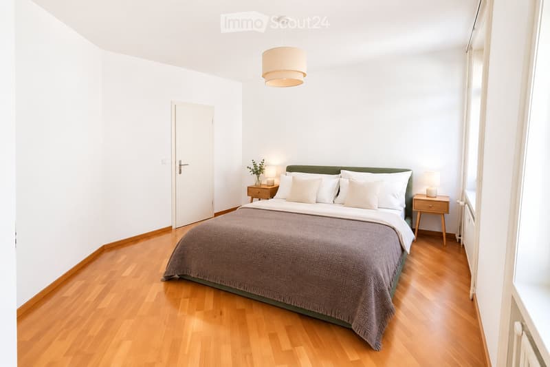 Minimalist bedroom with a wooden floor, white walls, a large bed with a green headboard, two nightstands, and a hanging pendant light.