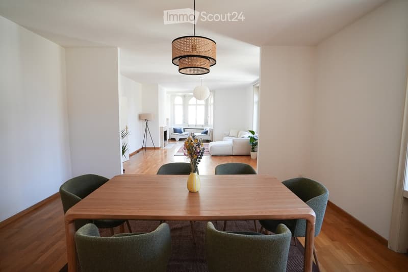 Bright, open living and dining area with wooden flooring, large windows, a dining table with six chairs, a sofa, an ottoman, a floor lamp, and decorative vases.