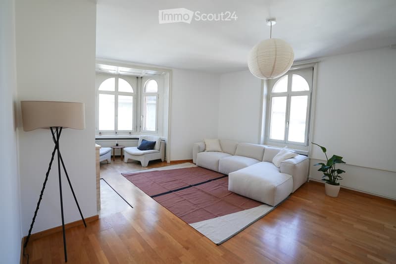 Bright living room with white walls, wooden flooring, large arched windows, a white sectional sofa with ottoman, two white armchairs, a small side table, a floor lamp, and a potted plant.