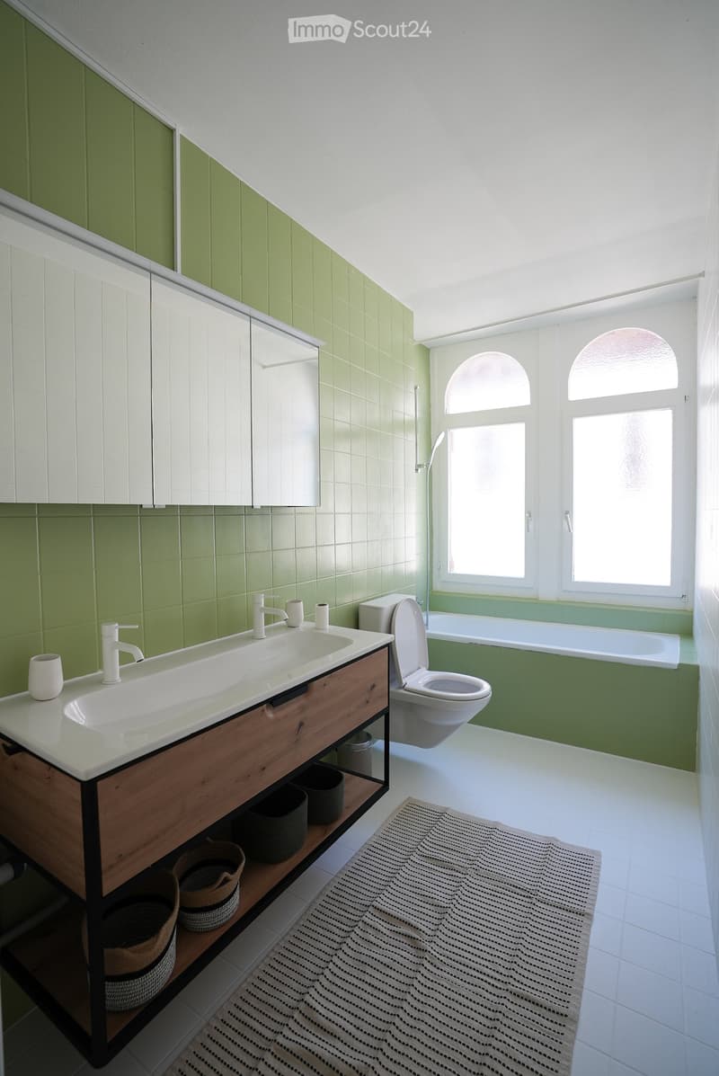 Modern bathroom with green tiled walls, white sink with wooden cabinet, toilet, bathtub, and large windows allowing natural light.