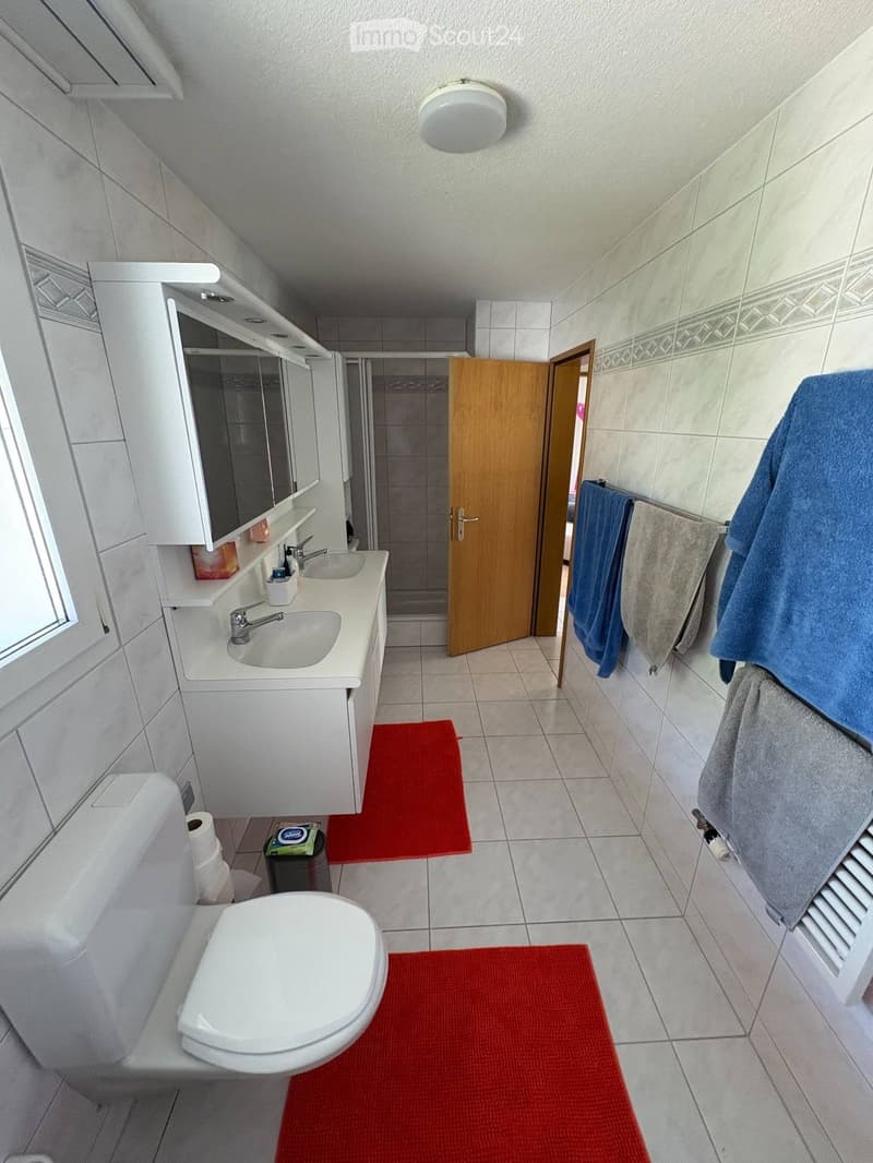 Modern bathroom with white tiles, a toilet, a sink with a mirror cabinet, and towels hanging on a rack. The floor has red bath mats.