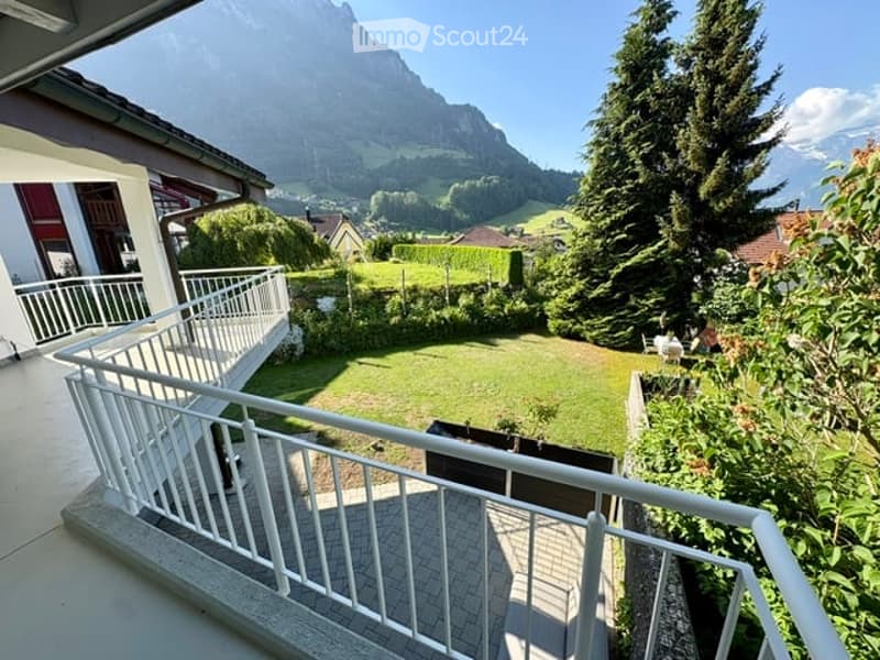 Balcony overlooking a green lawn and distant mountain range, with a white railing and tiled floor.